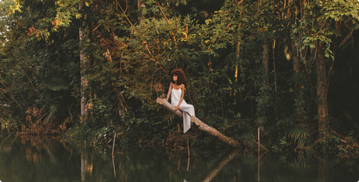 Mulher negra sentada em um tronco de árvore acima de um rio no meio da Amazônia olhando para o horizonte.